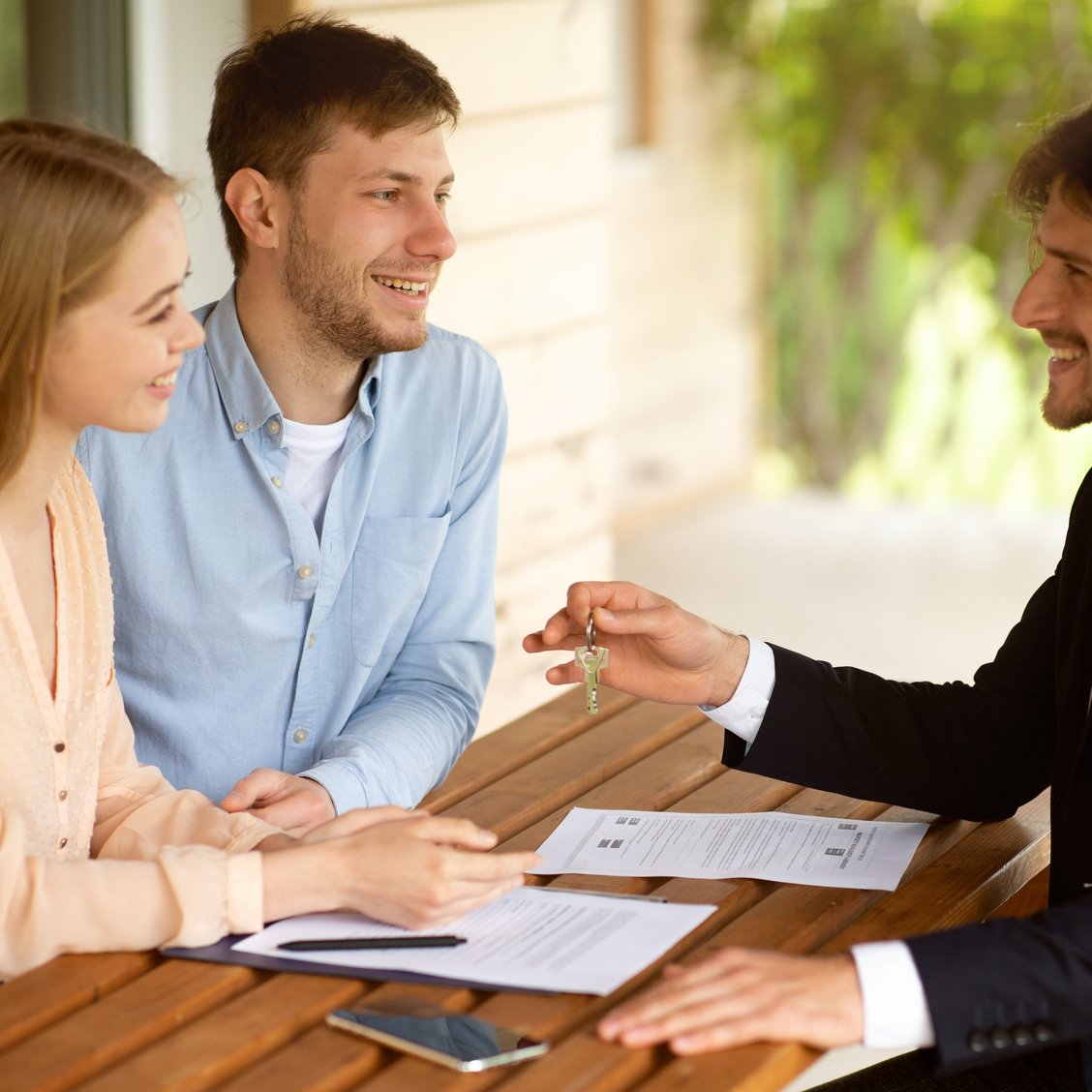 Real estate broker giving house key to millennial couple after signing property agreement at table outside Gestion locative Paris - Prestige investissement